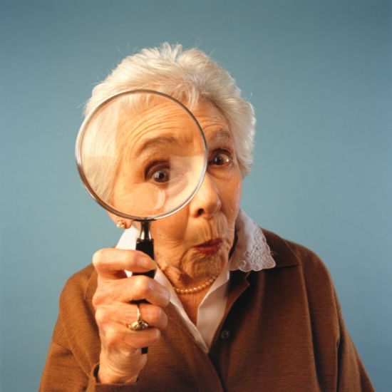 An older lady holds up a magnifying glass and looks directly at the reader.
