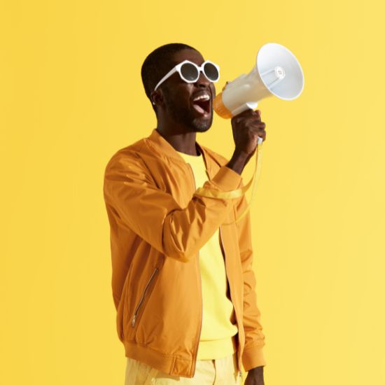 Man yelling into megaphone. He is dressed in a light yellow shirt, a dark yellow cardigan. The background is daisy yellow.